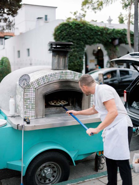 a pizza chef putting a pizza into a woodfired oven at Collen & Christian's Wedding Reception in the courtyard at Frequency Wine Company in Santa Barbara, California